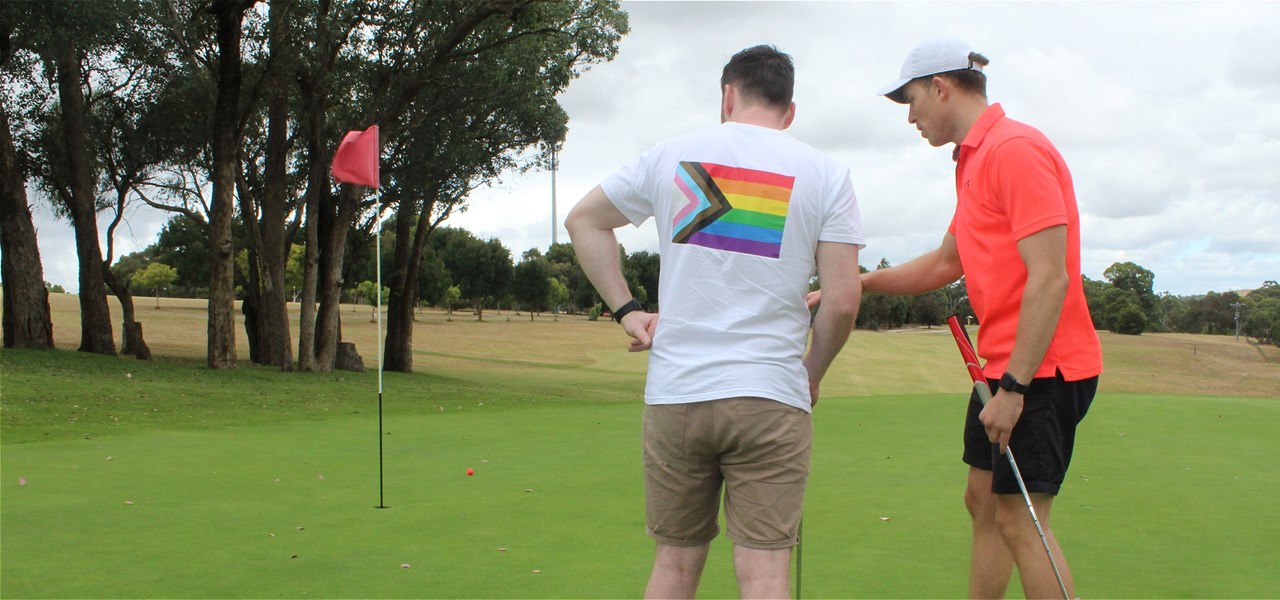 2 people are standing on a golf green looking at a red flag and are standing with their backs to camera