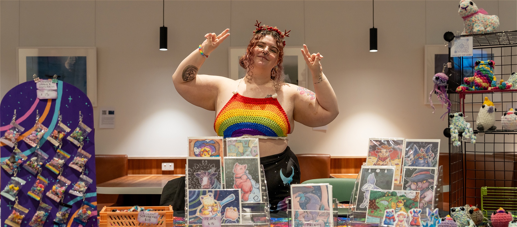 A queer person making a peace sign gesture stands behind their market stall of illustrations and amigurumi crochet merchandise.