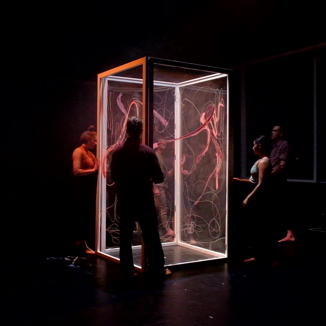 Glass cubicle on stage with performers surrounding it, audience in background
