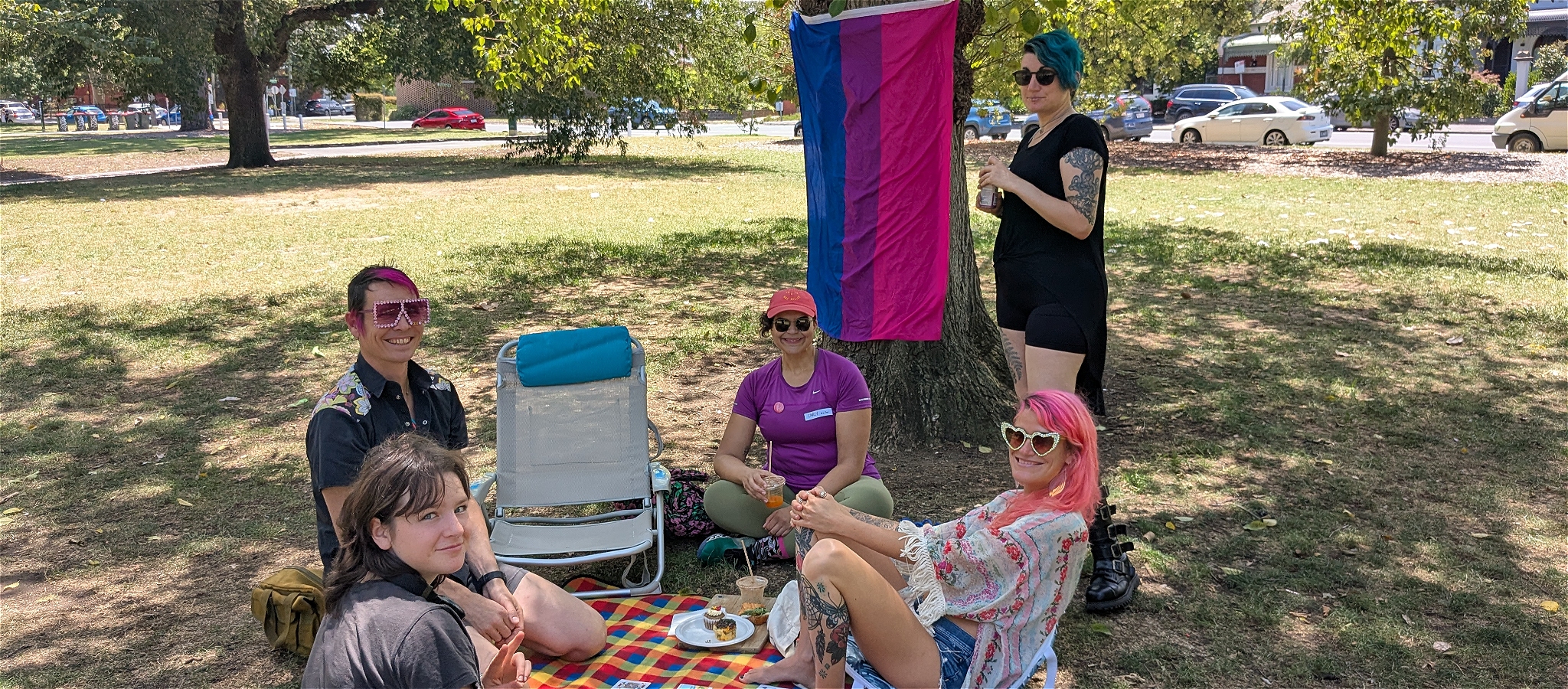 A group of smiling people with brightly coloured hair and clothes, sitting around a picnic rug with a bisexual flag behind them.