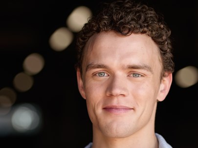 Ben is smiling with mouth closed. He wear a light blue linen shirt unbuttoned to show a white t-shirt underneath. He has blue eyes and short light brown hair with ringlet curls.