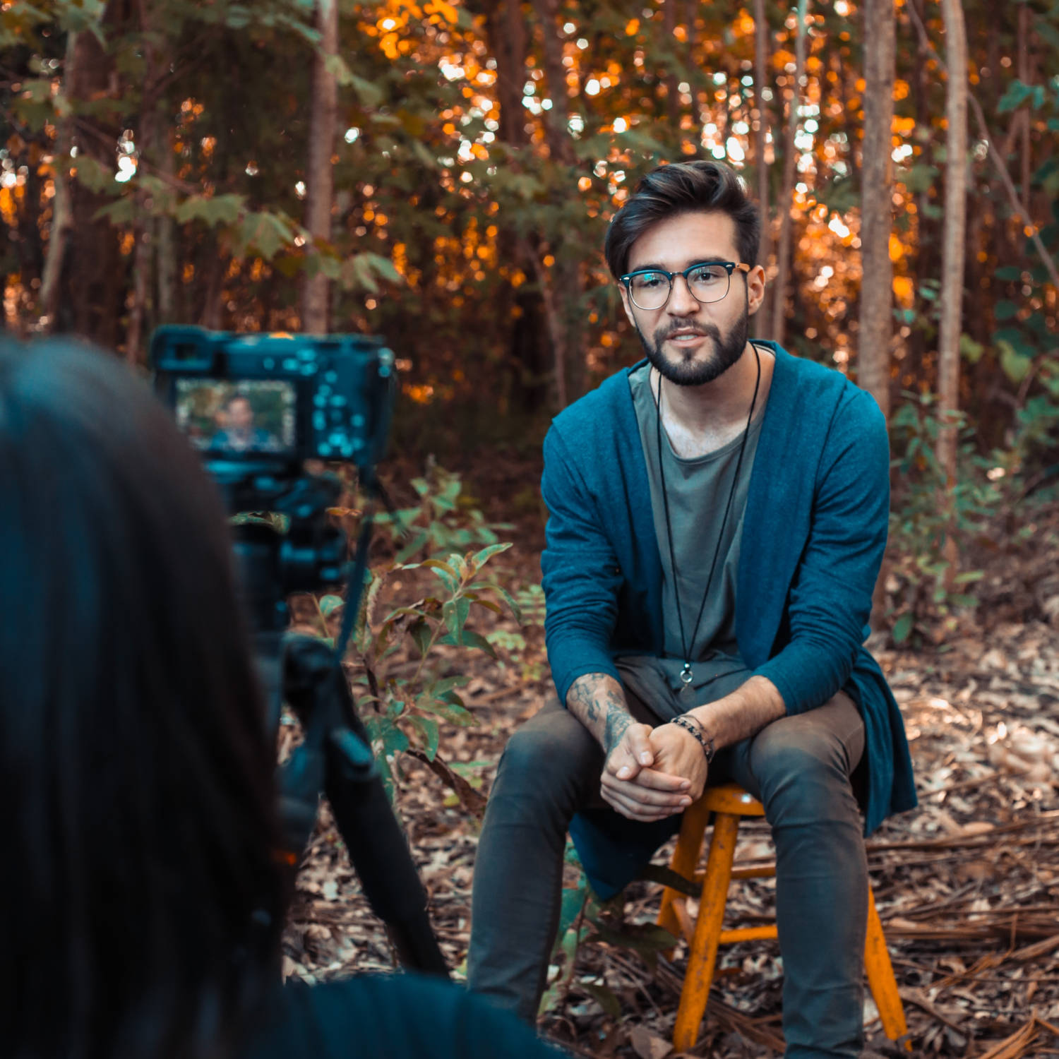 Seated man being photographed in a forest; photographer partly visible