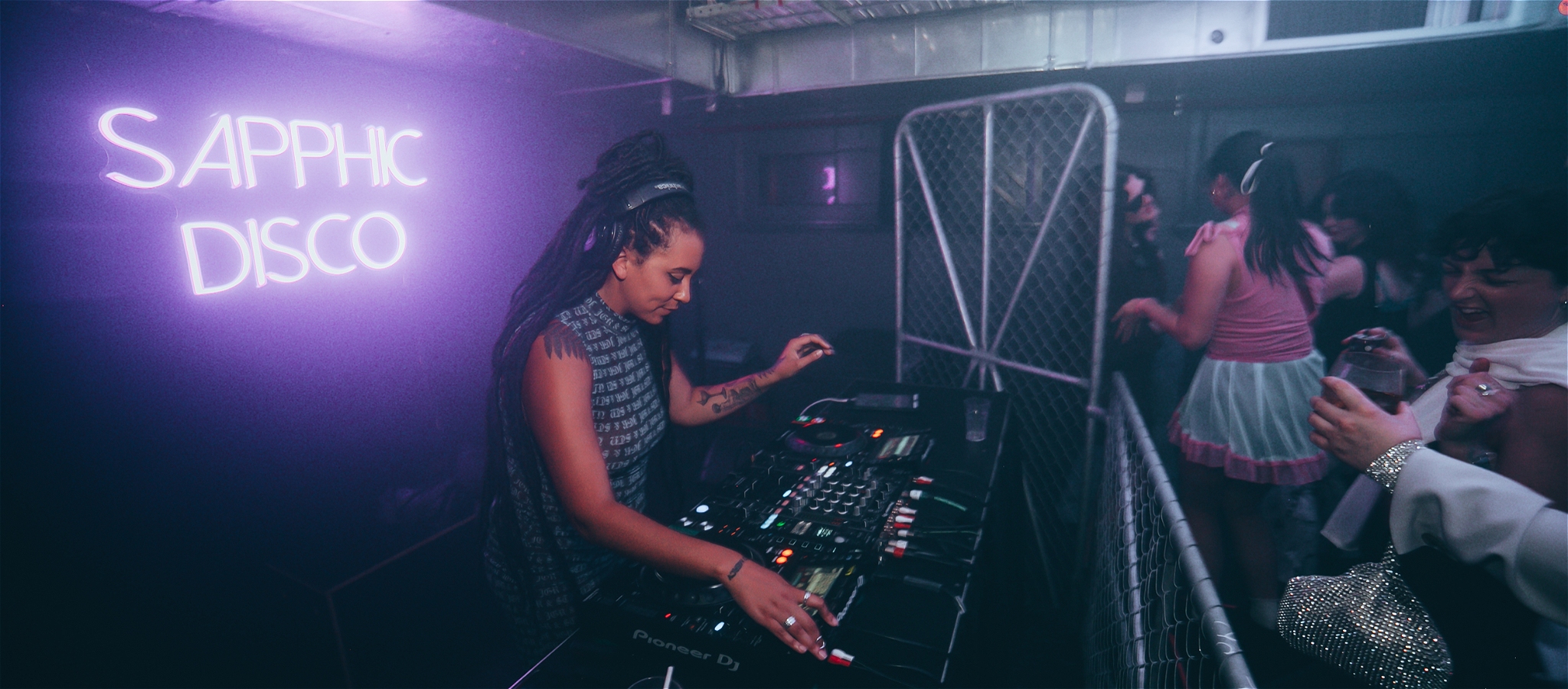 A female DJ plays music on decks backed by a neon sign that reads Sapphic Disco. In front of her people are dancing and singing.