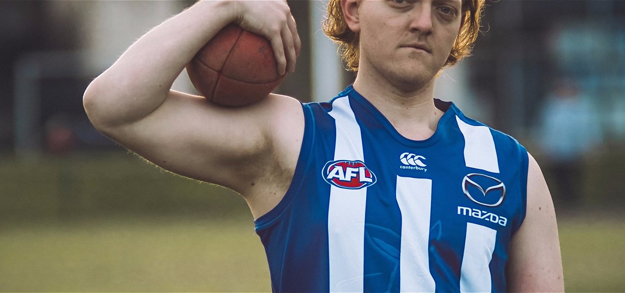 Blonde man wearing a blue and white AFL jersey stands on a field holding a ball. Their face is partially cut off in the photo.