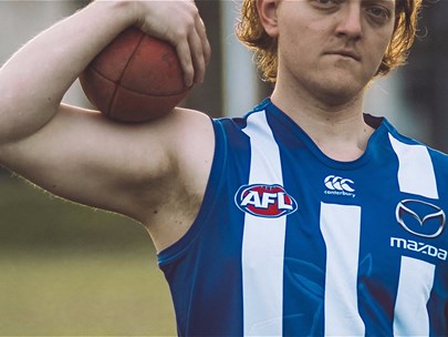 Blonde man wearing a blue and white AFL jersey stands on a field holding a ball. Their face is partially cut off in the photo.