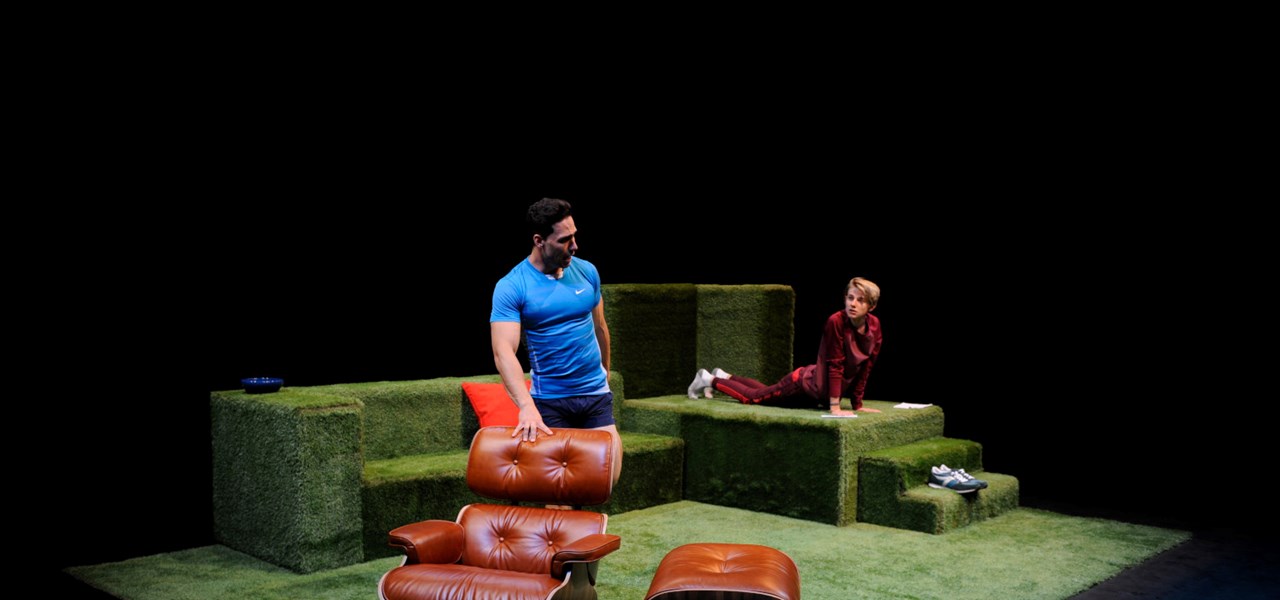 Two actors on stage: one in a blue t-shirt standing behind a recliner chair, the other doing press-ups in the background