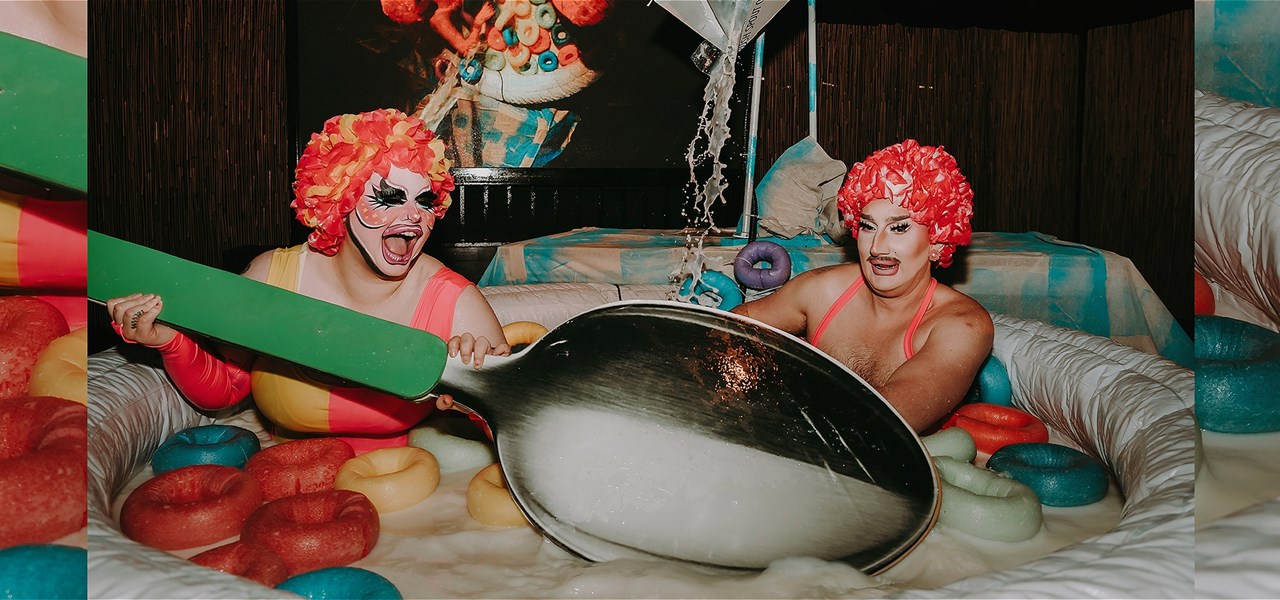 A drag queen holding a giant green wooden spoon in a human sized milk bowl with another drag queen playing in giant cereals.