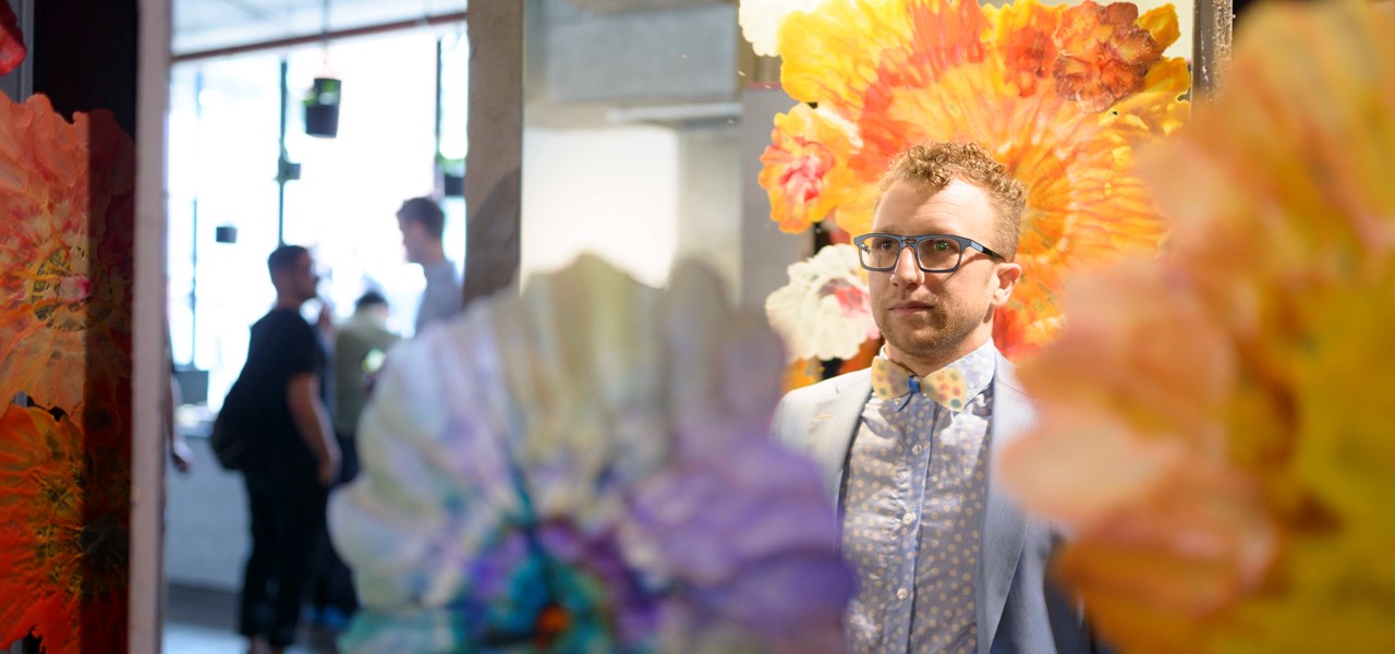 Elegantly dressed man wearing glasses in an art gallery, surrounded by colourful umbrellas and feathers