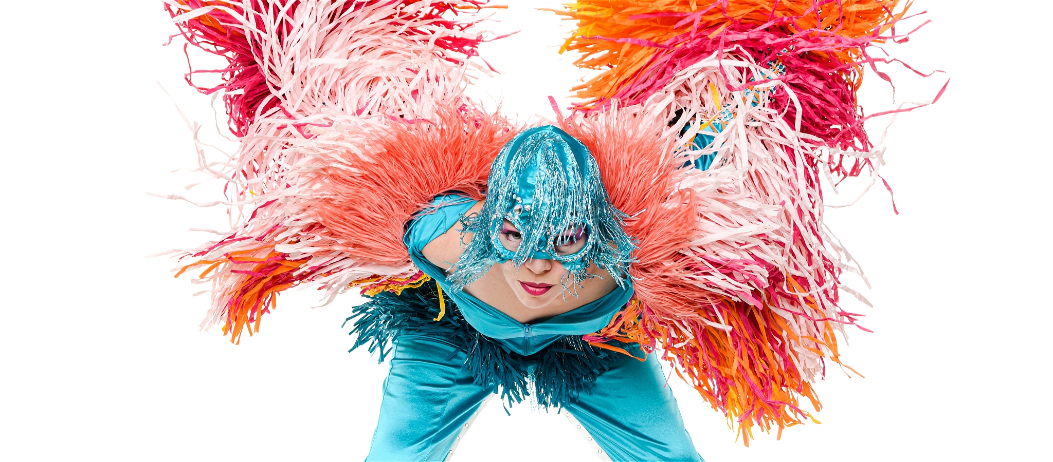 Imogen Kelly is in a turquoise satin superhero suit with huge pink cerise orange and yellow tassels on the arms and legs.