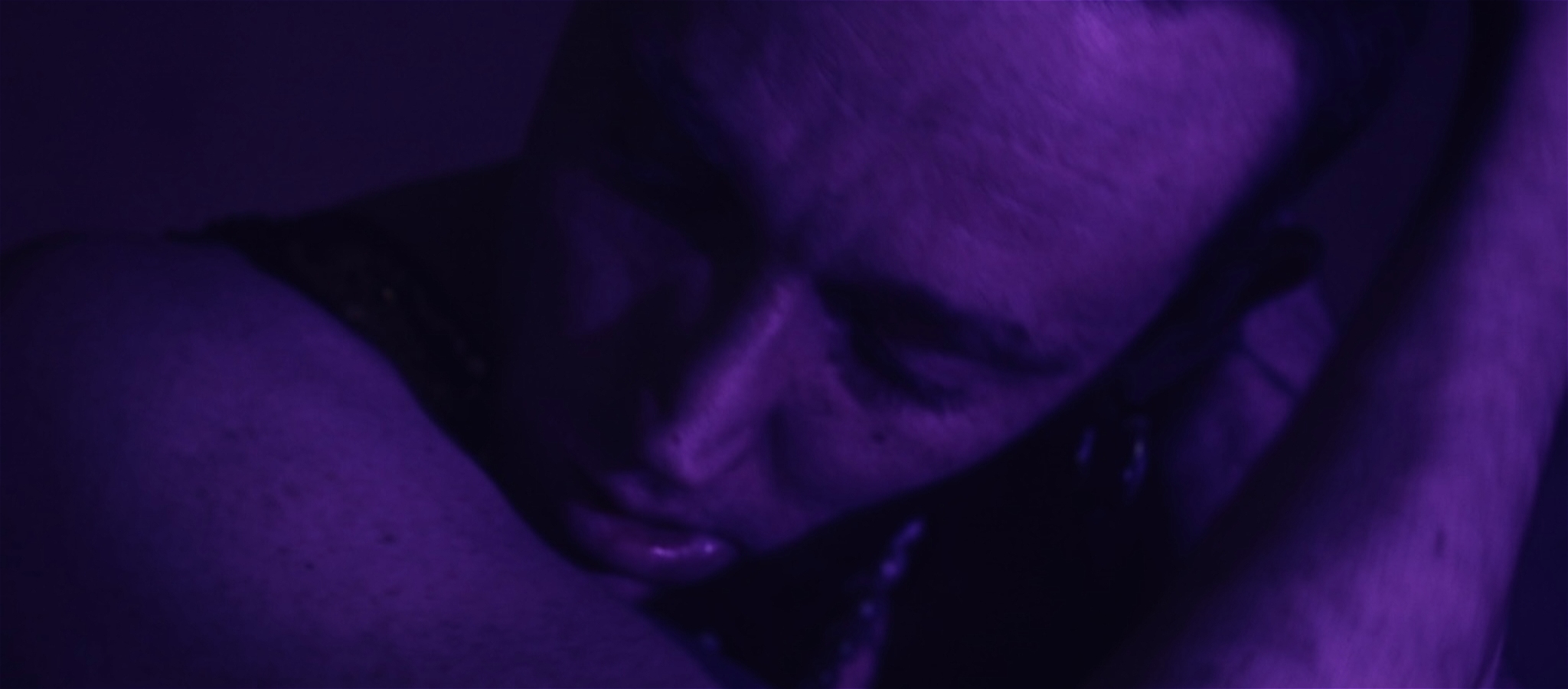 A close up shot of a non binary person with eyes closed looking down- the image is deep purple