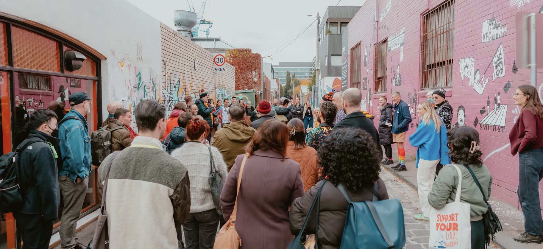 A crowd of about 30 people stand on a cobblestone street amongst eclectic Collingwood buildings. The building on the right features the large, pink 'Retracing Queer Footprints' mural