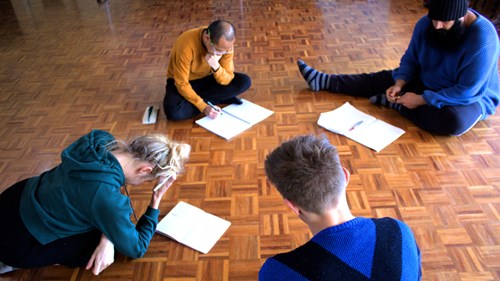 Four people sitting on a parquetry floor with pen and paper, looking pensive