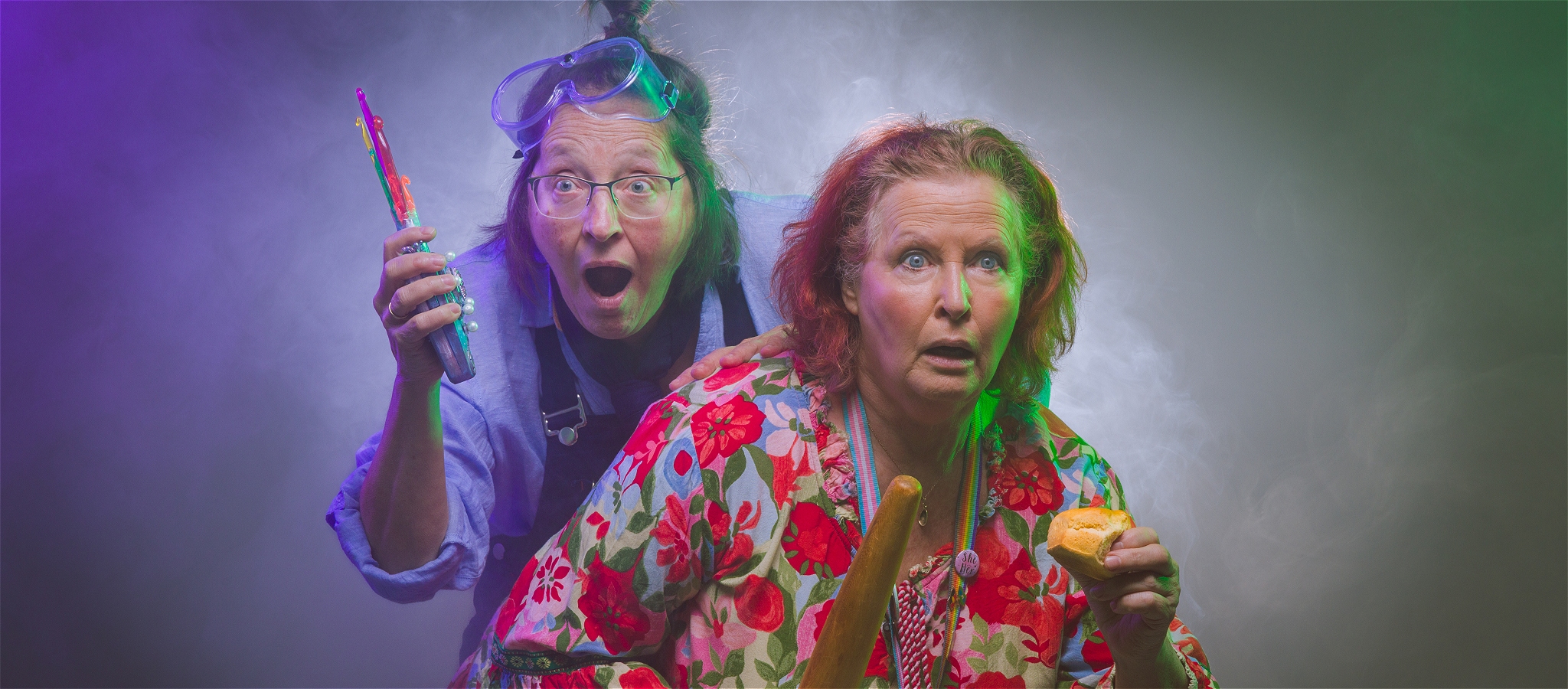 Two older women with shocked expressions on their faces holding baked goods and knitting needles.