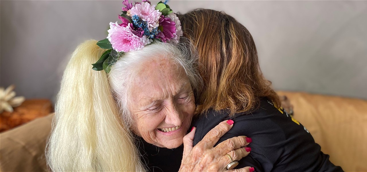 Older women hugs someone on a couch, clutching their shoulder with a smile.