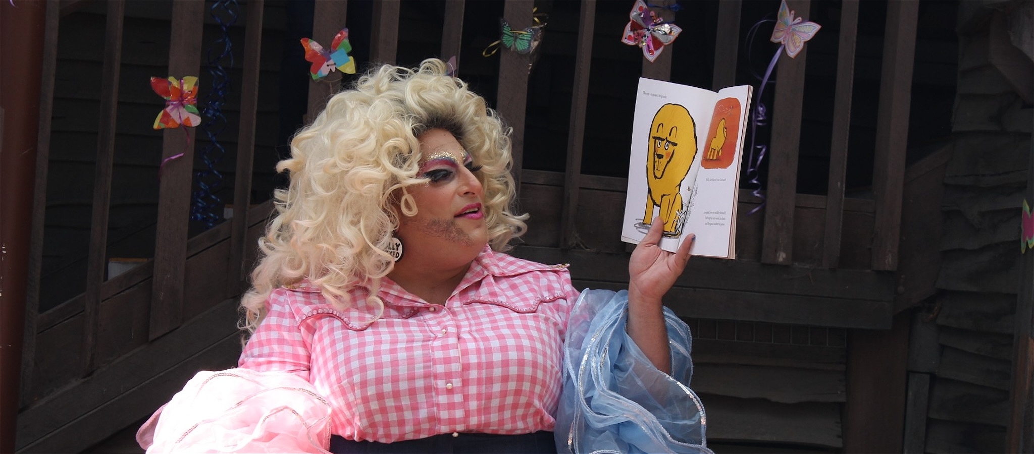 A person with blonde hair wearing colourful makeup, a pink and white checked dress holding up a story book they are reading.
