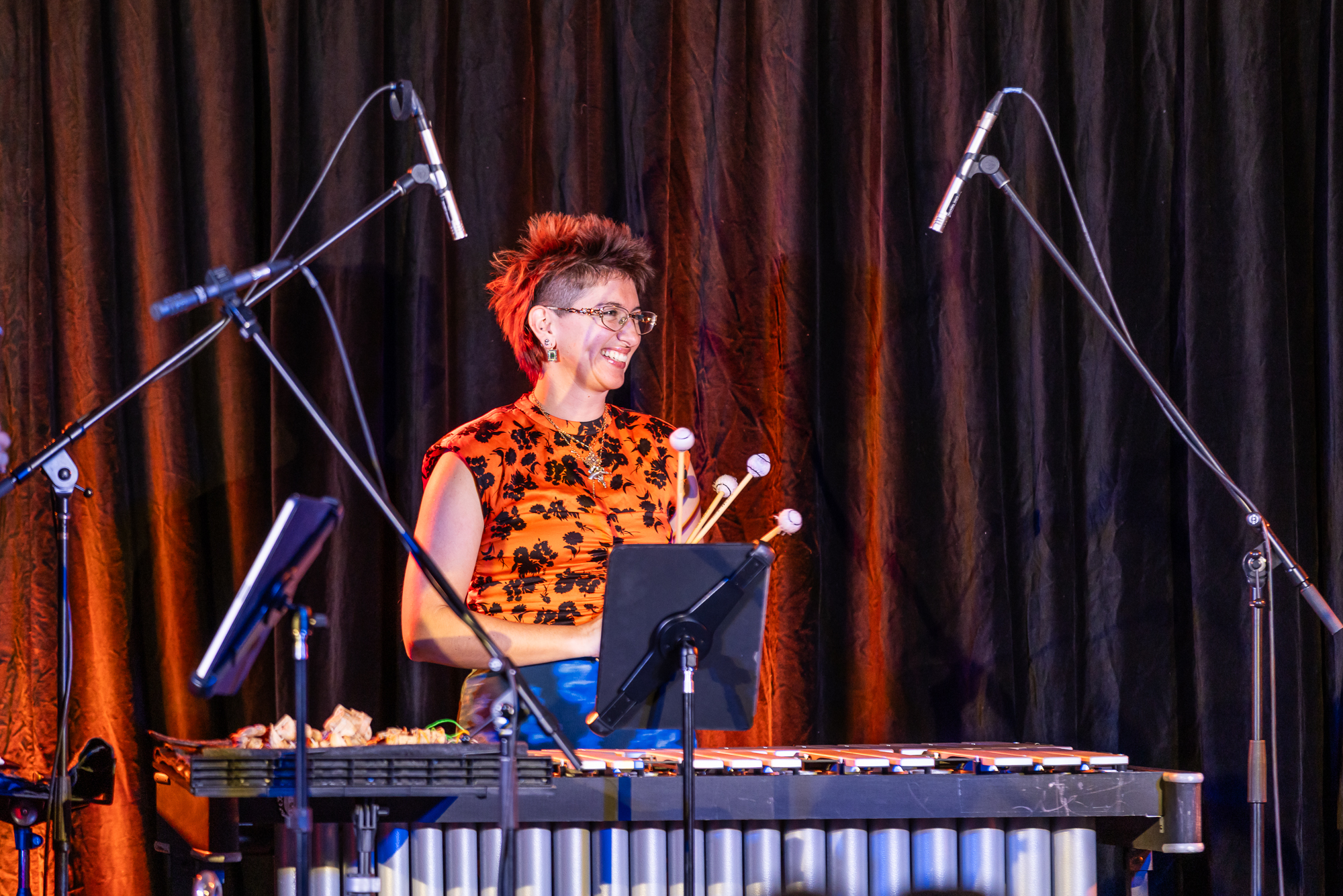 A smiling Niki Johnson wearing a bright orange short sleeve top with a black pattern on it, stands behind a long flat percussive instrument holding beaters. Niki stands infront of a curtain and is lit by a spotlight. There are many microphones surrounding her to record her performance.