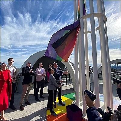 An open rooftop. A group of people are watching an LGBT flag being raised.