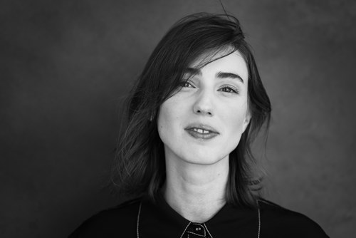 A black-and-white photograph of Amy, half smiling in front of a neutral background with their dark hair partially covering their eyes. 