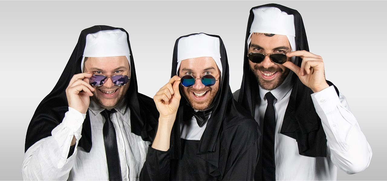 Three men dressed as nuns, styled with white shirts and black ties, smile into the camera while holding sunglasses below their eyes.