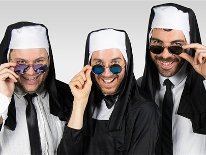 Three men dressed as nuns, styled with white shirts and black ties, smile into the camera while holding sunglasses below their eyes.