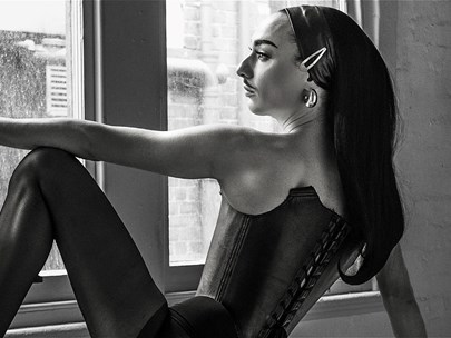Black and white photo of a dancer-like person with long black hair seated and looking pensively out a window