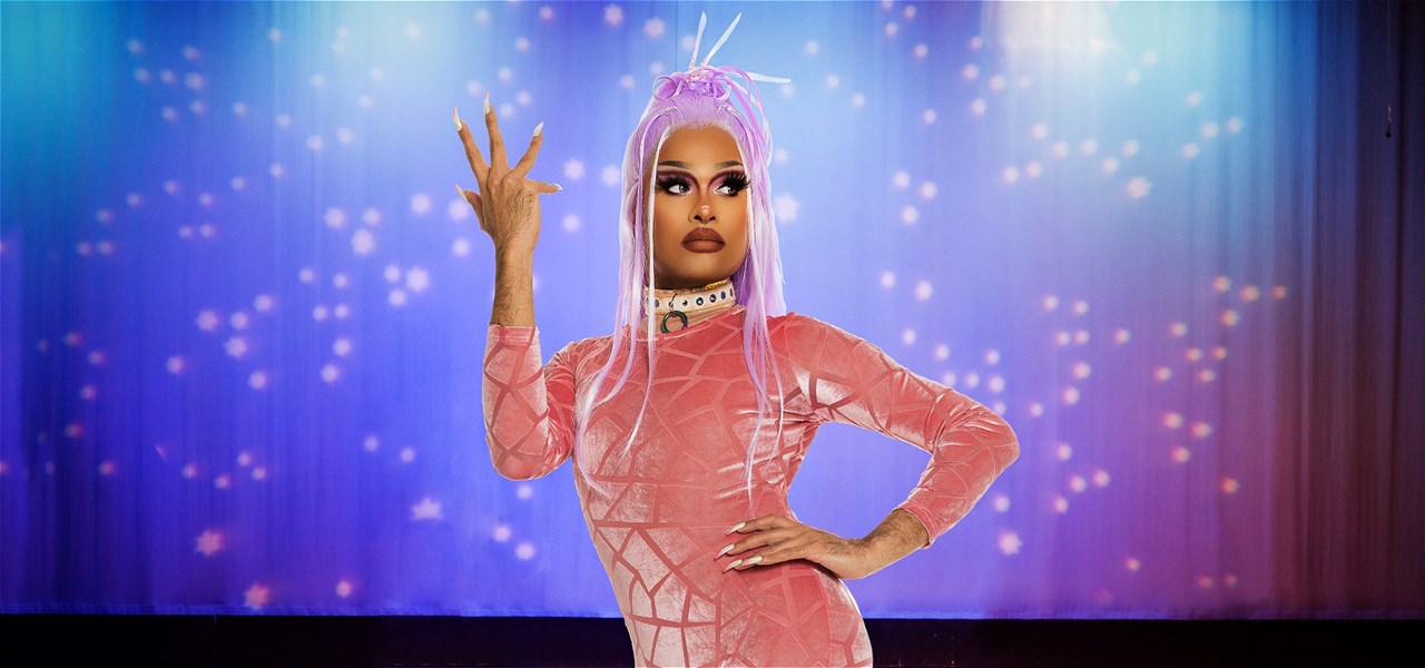 Drag queen Cerulean wearing a purple wig and orange catsuit, standing in front of a stage.