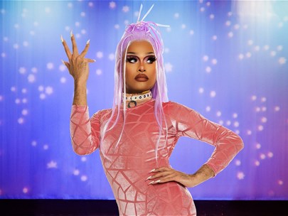 Drag queen Cerulean wearing a purple wig and orange catsuit, standing in front of a stage.