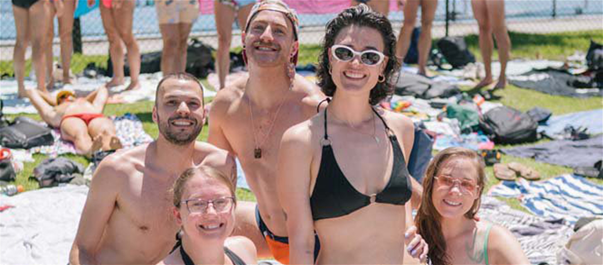Five people sitting on the grass in the sun outside and smiling looking at the camera.