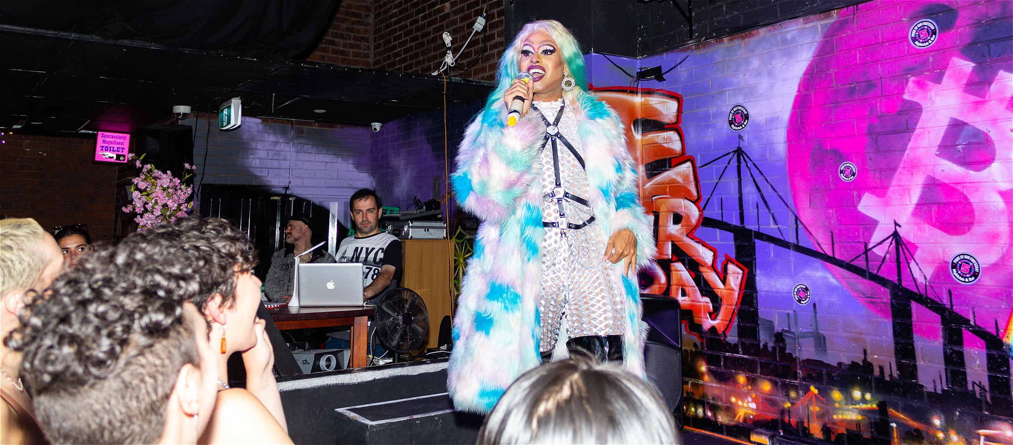 A drag queen wearing a multicoloured fur coat stands on stage in front of an audience with microphone raised to her mouth.