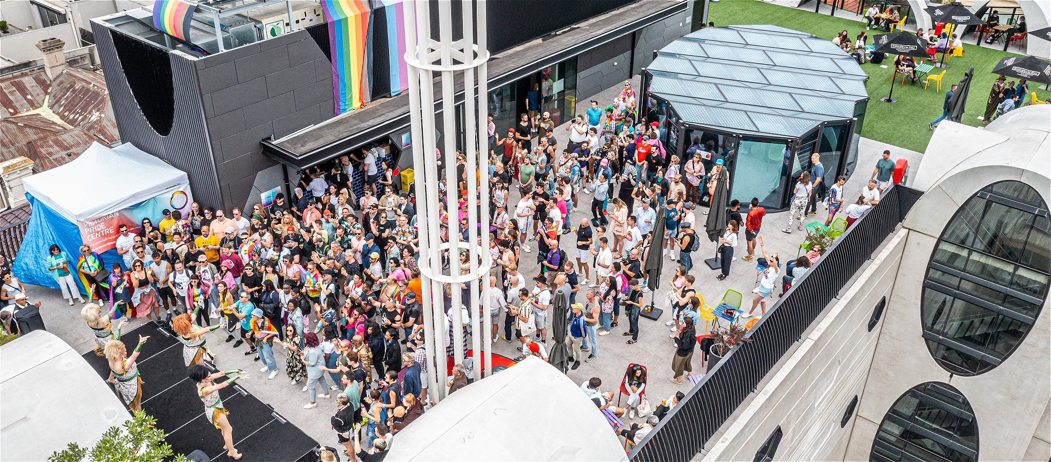 An image of a rooftop party with a stage and some drag queens in front of a large crowd