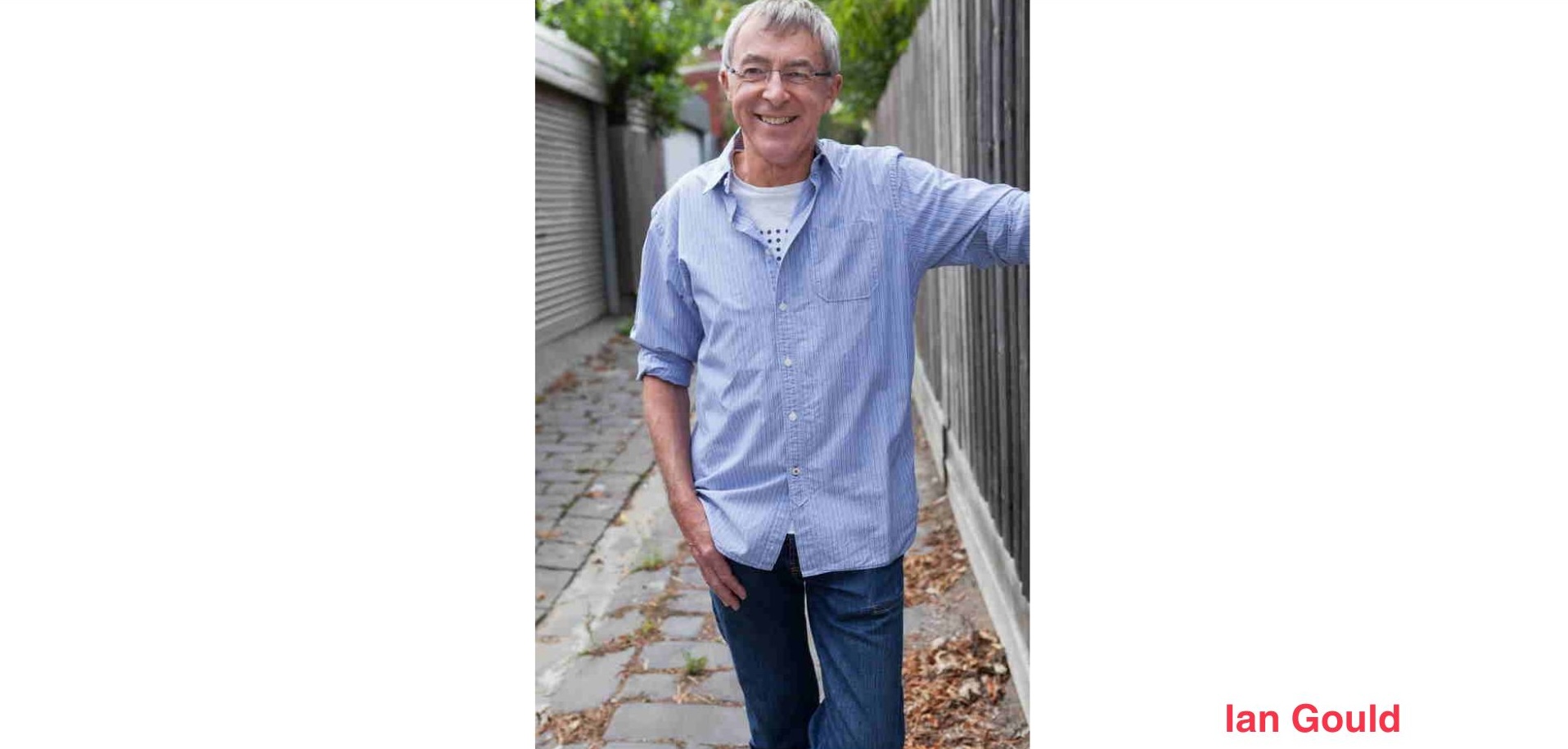 Ian Gould standing in a laneway smiling while dressed in jeans and a blue shirt