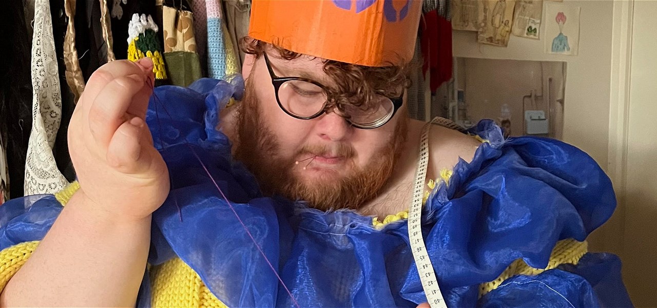 Male-identifying person wearing a blue and yellow dress while sewing something by hand