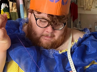 Male-identifying person wearing a blue and yellow dress while sewing something by hand