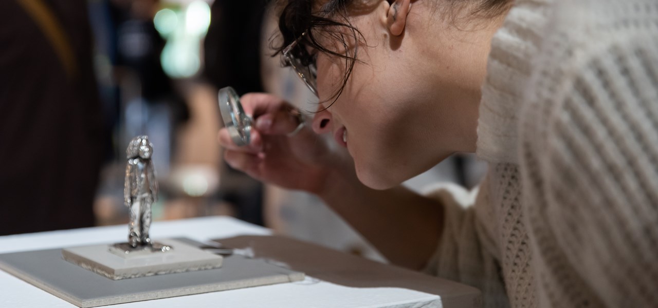 A person with a magnifying glass leaning in to look at a small solid silver sculpture
