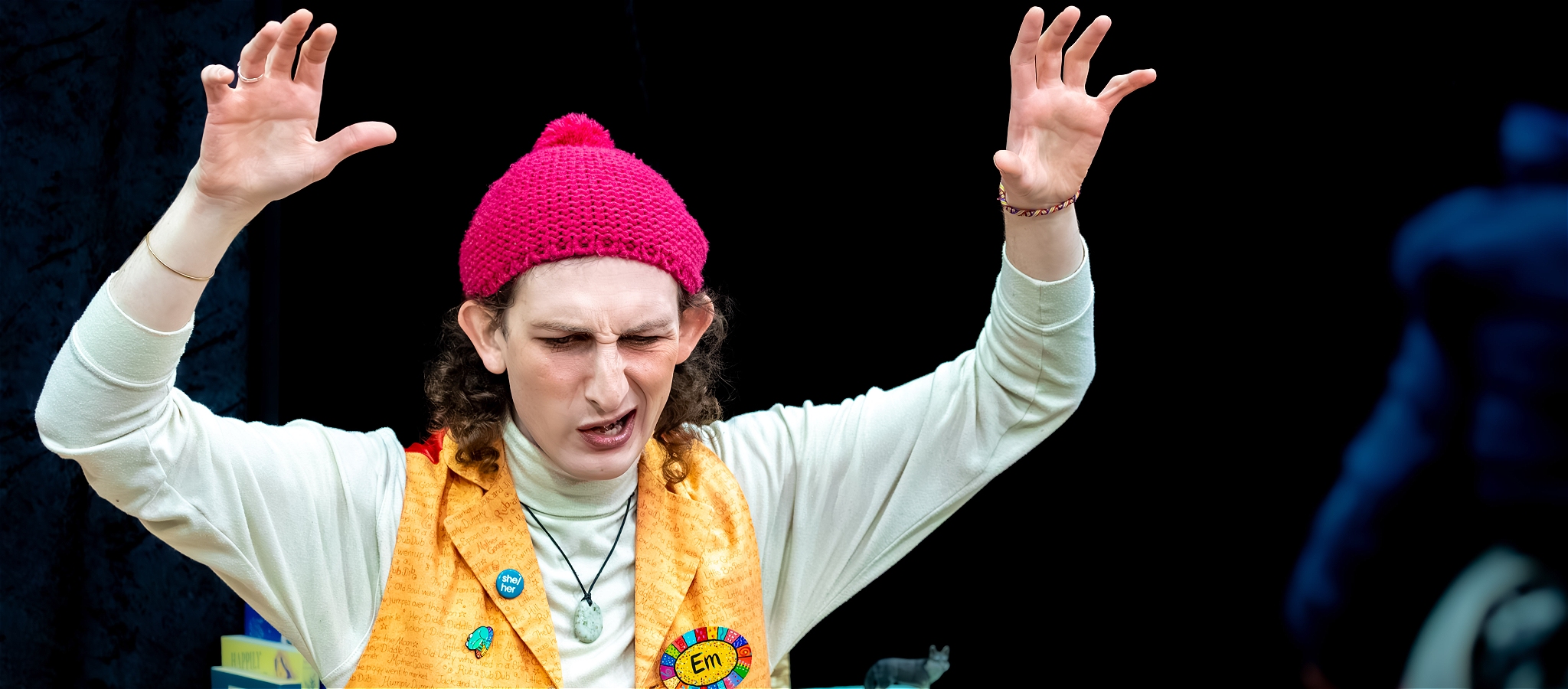 A person wearing a pink hat, white top and yellow vest with their hands up, making a grimace face