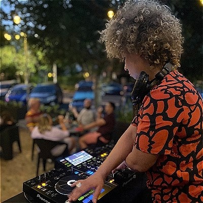 DJ in the foreground with people sitting at tables in the background then parked cars in the street behind them