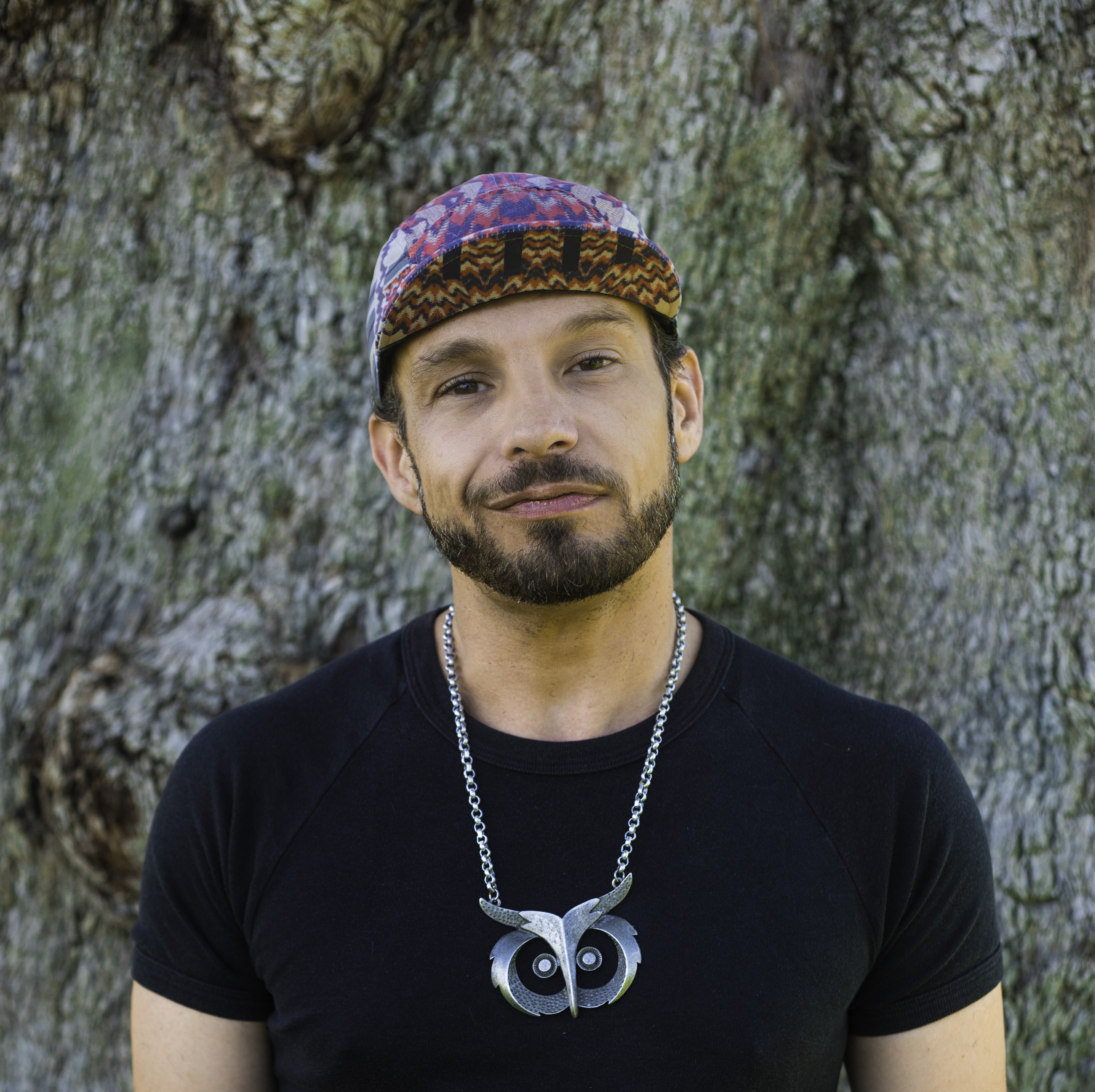 a person with dark brown hair and a beard wearing a colourful cap and an neckalace.