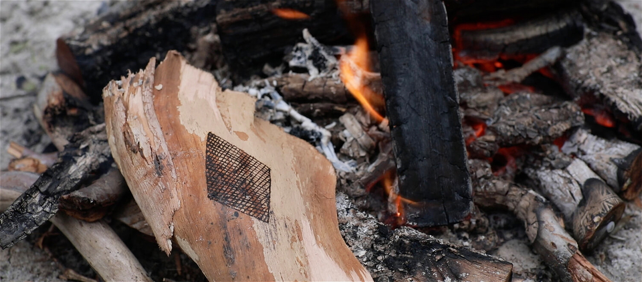 A diamond shaped pattern with cross hatching etched into a piece of wood placed within a bonfire with flames and charred sticks