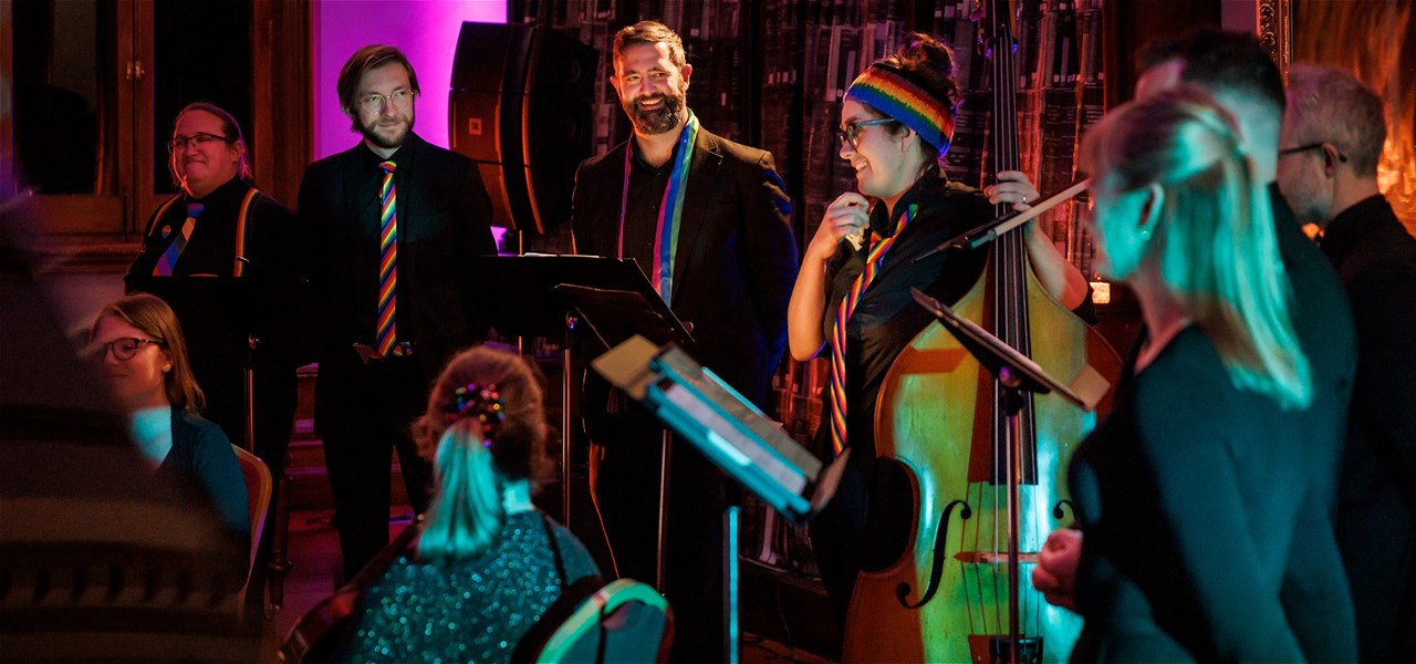 Singers and string players stand on a stage, the double bass player is talking and laughing
