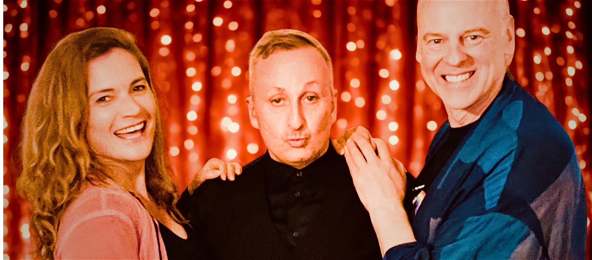 Three cabaret artists standing on stage smiling in front of a red curtain with fairy lights.