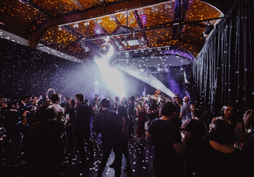 Crowd standing looking at a stage, with several spot lights on the stage, and glittering lights around the room