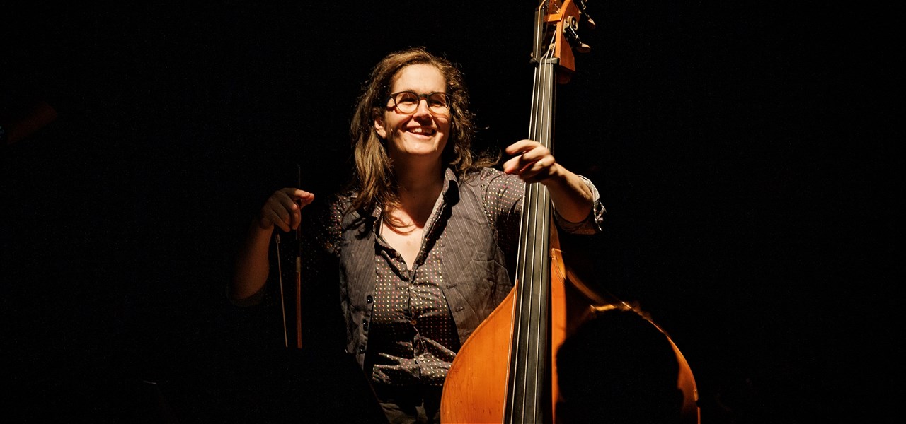 Long haired butch with glasses, white woman with brown hair, stands holding a double bass and smiling