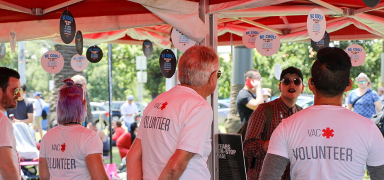 View looking out from a VAC stall at Midsumma Carnival, with all stallholders wearing white t-shirts saying VOLUNTEER on the back