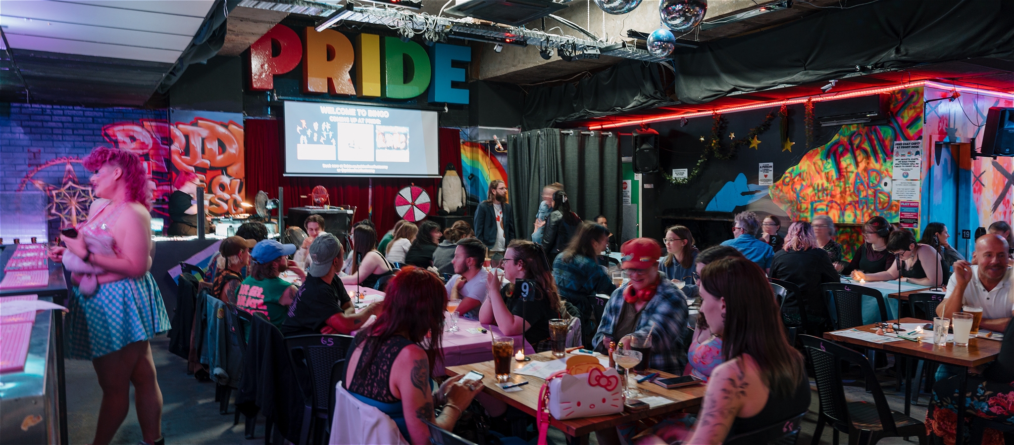 A crowded room of people at tables, ready to play bingo. On the far left of the image, a brightly dressed person waits at the bar.