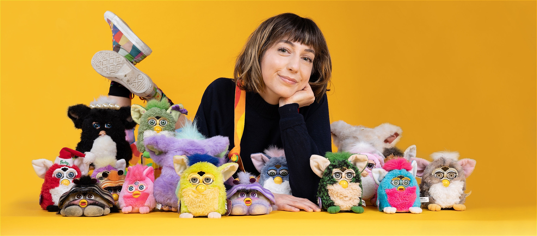 A smiling person lying on the ground amongst a group of Furbys of different colours, sizes and designs with a yellow background.