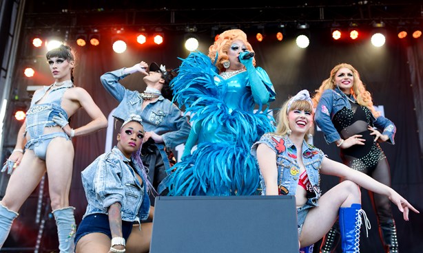 Drag queen singing, surrounded by elegantly dressed show people; all costumes are blue
