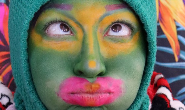 Headshot of a person whose head is wrapped in a beany-like headdress. Their face is heavily painted and there's a colourful, abstract background.