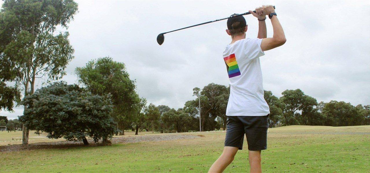 A person wearing a white t-shirt with a pride flag printed on the back is standing on a golf course with their golf club lifted, preparing to swing