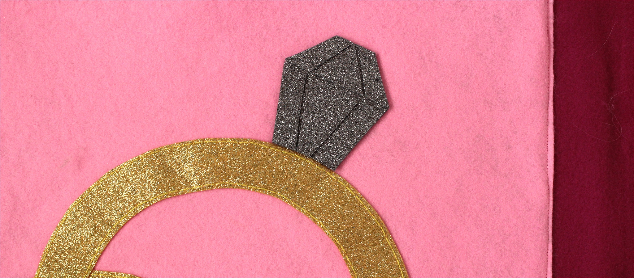 An image zoomed in of one of Tess Hiders fabric works. The viewer can see a gold circular ring with a grey diamond shape, and a light pink background