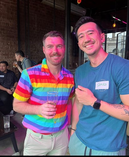 Two male-identifying people smiling for the camera. One in a rainbow shirt the other a grey t-shirt.